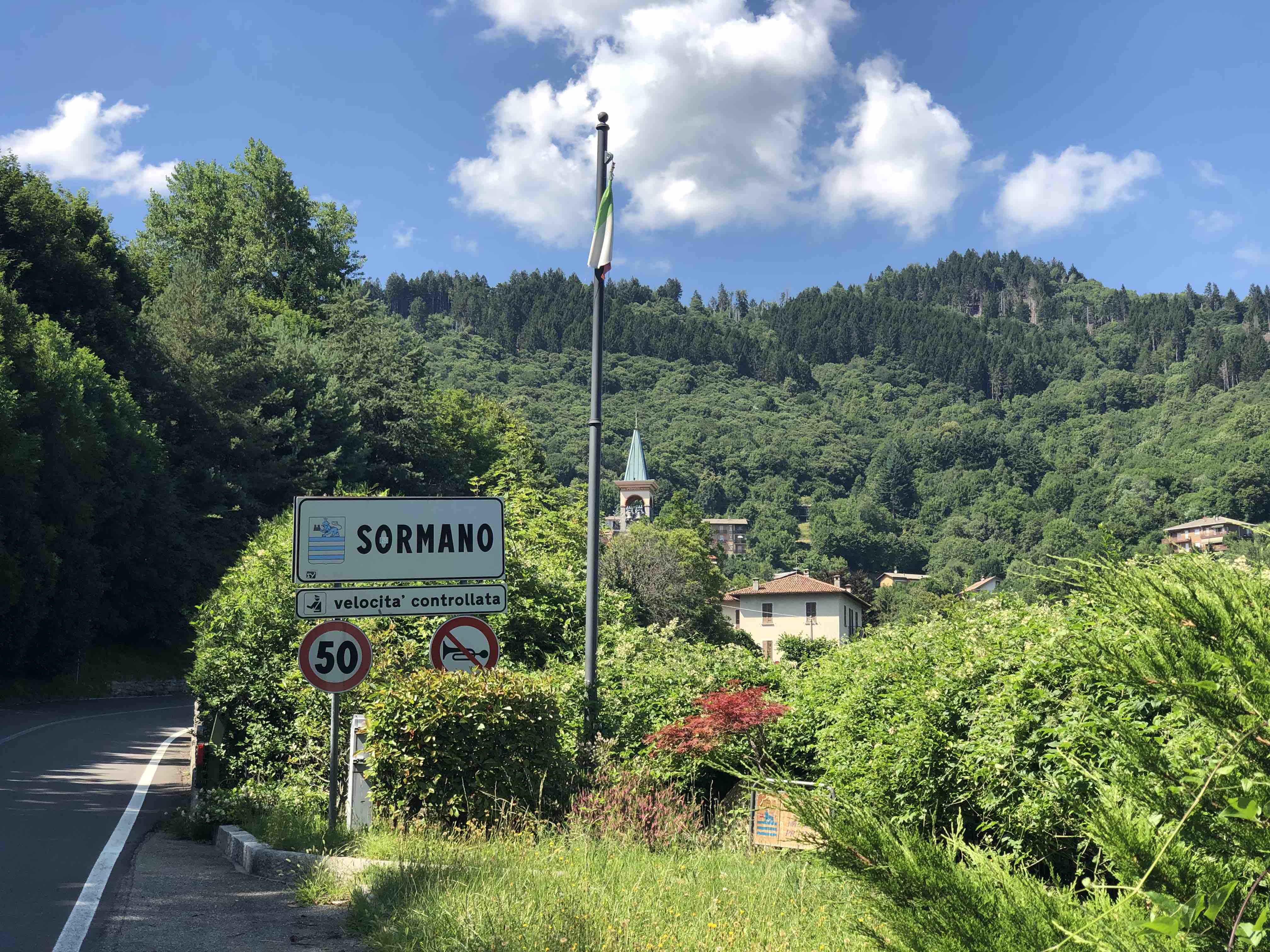 View out over the town of Sormano, Italy with flag pole, green trees, and blue sky with church in the background