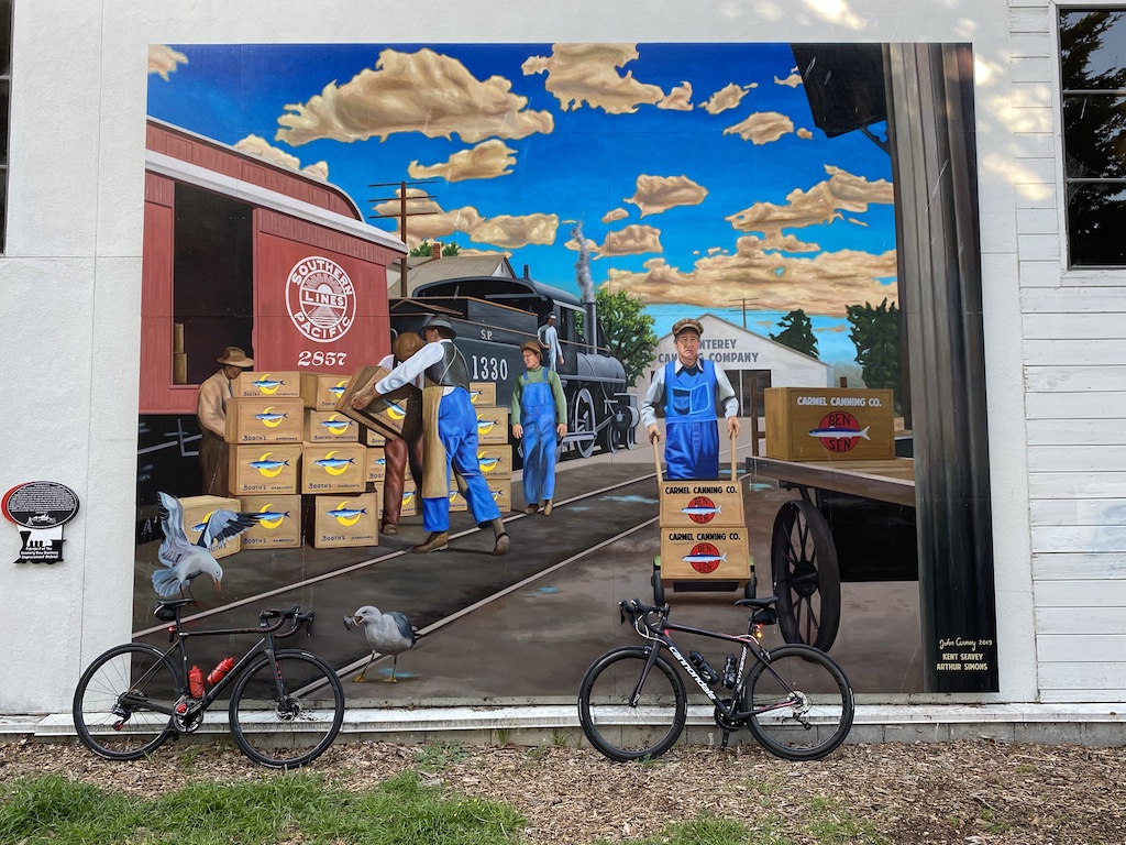 Factor O2 VAM and Cannondale Synapse bikes resting against a mural along the bike path in Monterey, California depicting men unloading crates from a railroad car