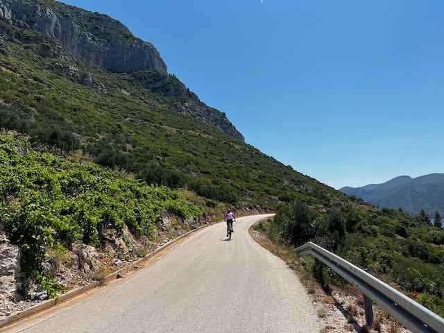 A coastal ridge road with a cyclist in front of a large rocky cliff face in the distance